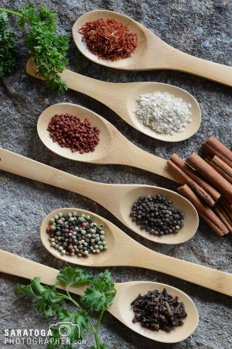 Overhead view of six wooden spoons each holding different spices with parsley and cinnamon accents