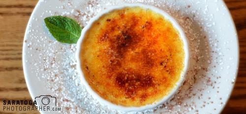 Overhead view of crème brûlée on white plate dusted with confectionery sugar and mint leaf accent