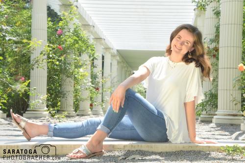 Young woman in white top with bluejeans seated and smiling at camera with pergola and Rosegarden behind her