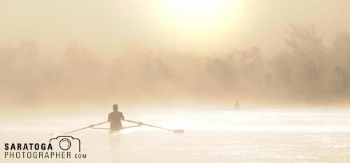 Man rowing single racing skull on water with fog and hazy sun in background